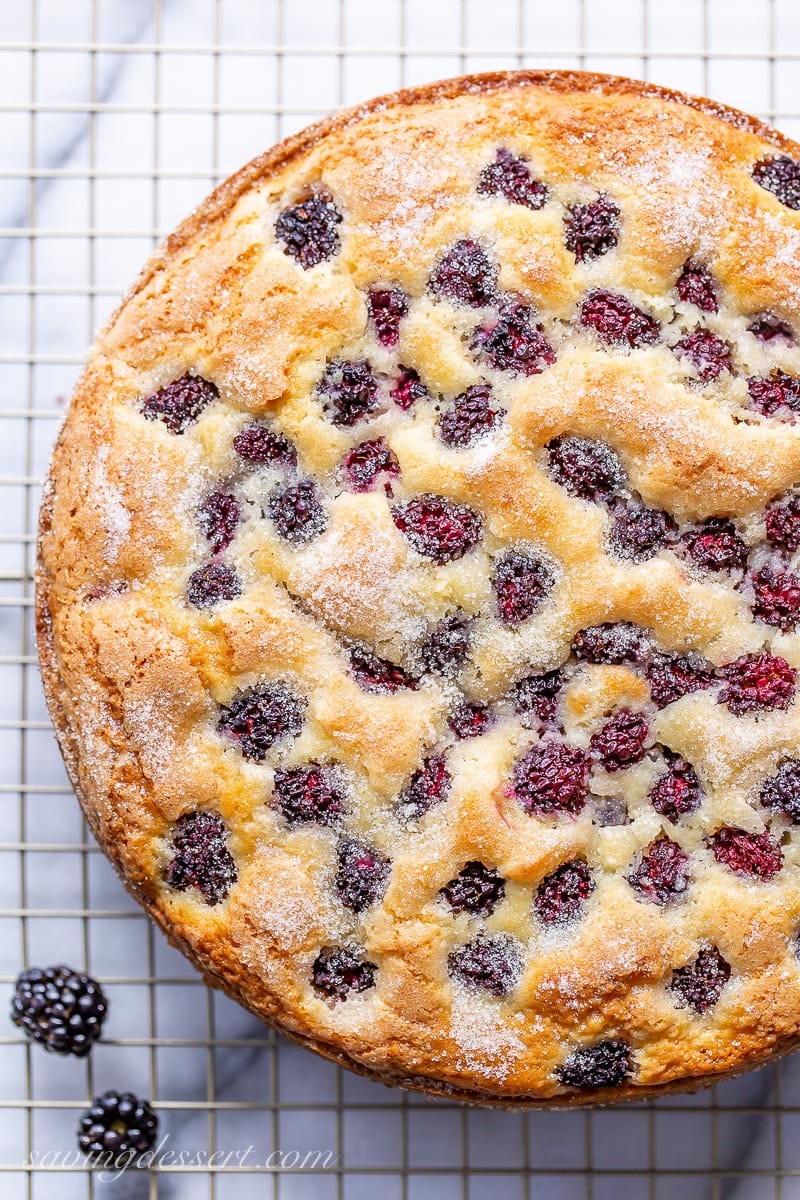 An overhead view of a breakfast cake with a sugary, crisp top and blackberries on a cooling rack