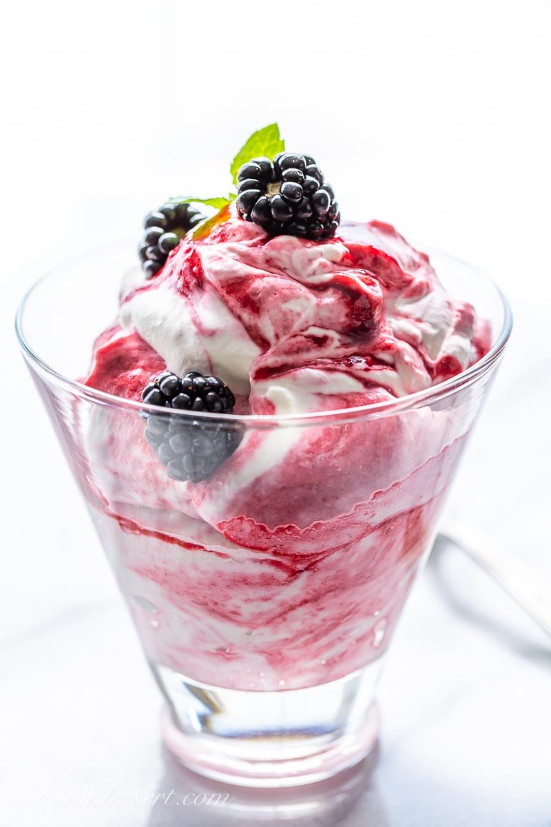 A clear glass filled with a blackberry swirled whipped cream topped with fresh blackberries and mint leaves