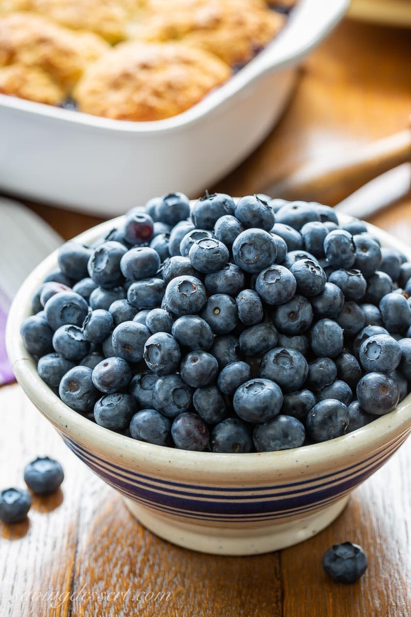 A bowl of fresh blueberries with a cobbler in the background