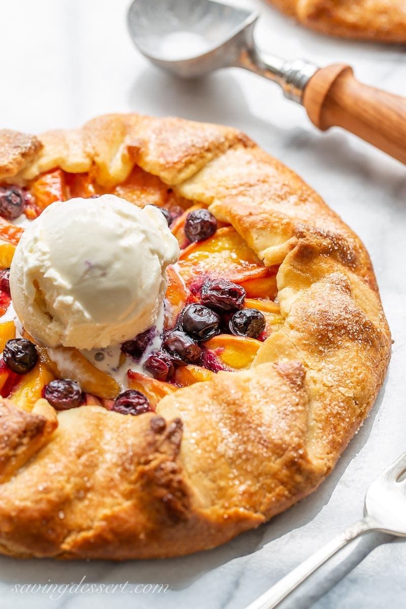 A golden brown peach galette topped with blueberries and a scoop of vanilla ice cream
