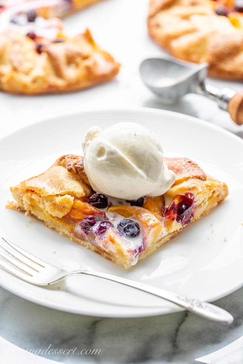 A slice of a baked galette with peaches and blueberries topped with a scoop of vanilla ice cream