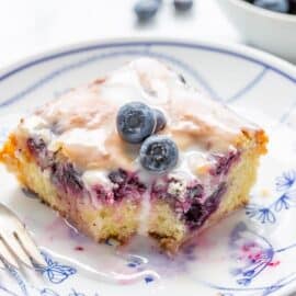 A slice of blueberry lemon zucchini cake on a plate topped with a few fresh blueberries and a lemon glaze
