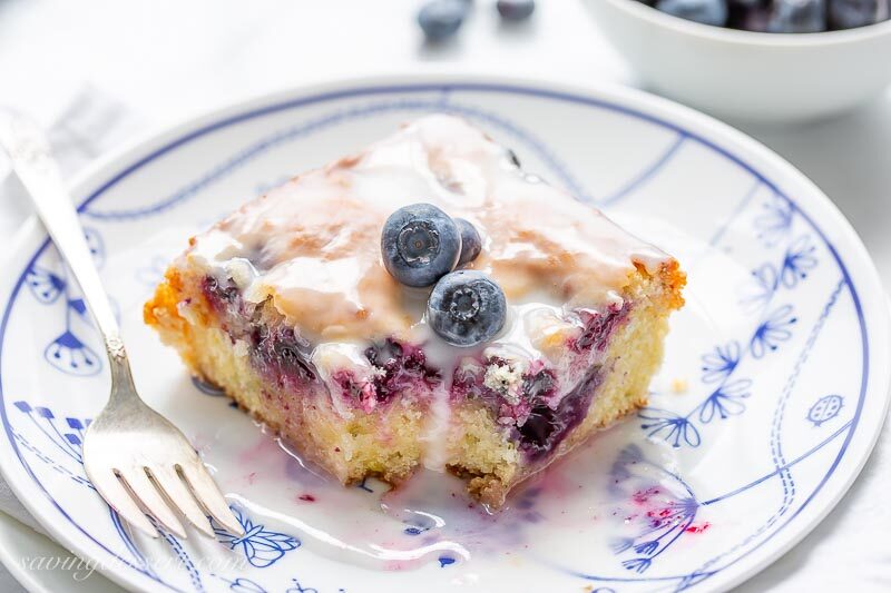 A slice of blueberry lemon zucchini cake on a plate topped with a few fresh blueberries and a lemon glaze