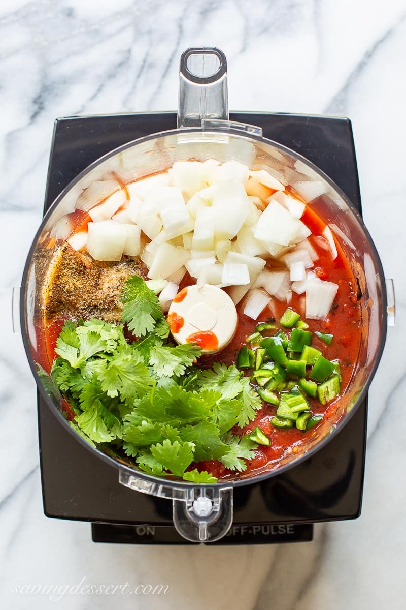 A food processor bowl filled with tomatoes, cilantro, jalapeños, spices and diced onion