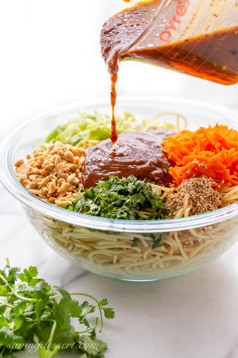 A bowl of spaghetti, peanuts, carrots, green onions and cilantro being drizzled with a spicy peanut sauce
