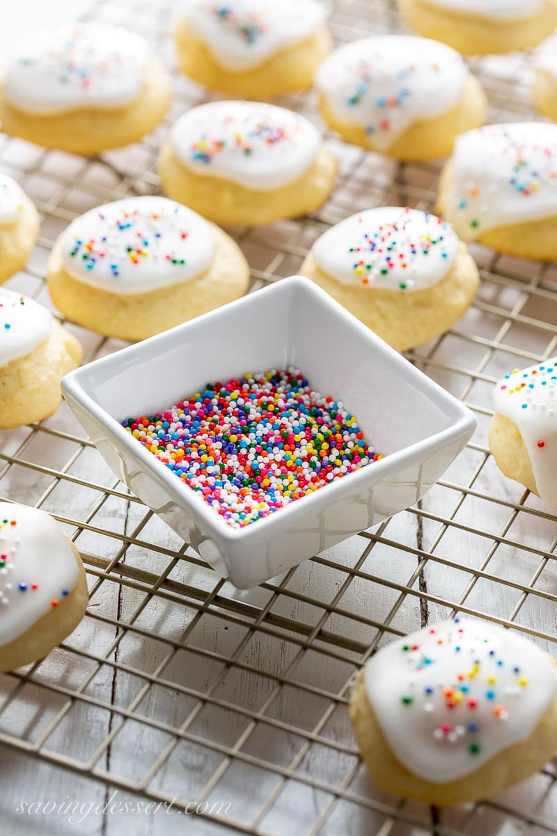 A bowl of sprinkles and a cooling rack filled with soft Ricotta Cookies with a simple glaze on top