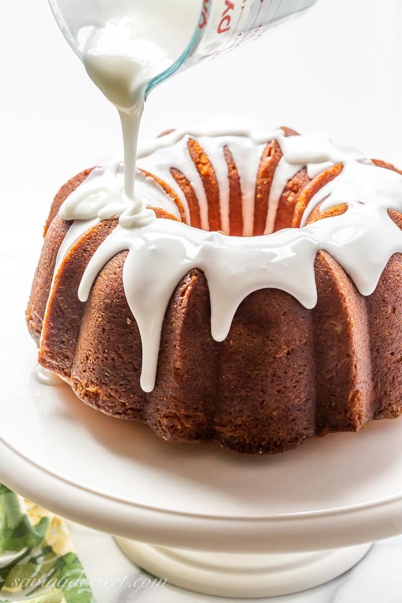 A lemon bundt cake being drizzled with a lemon icing from a measuring cup