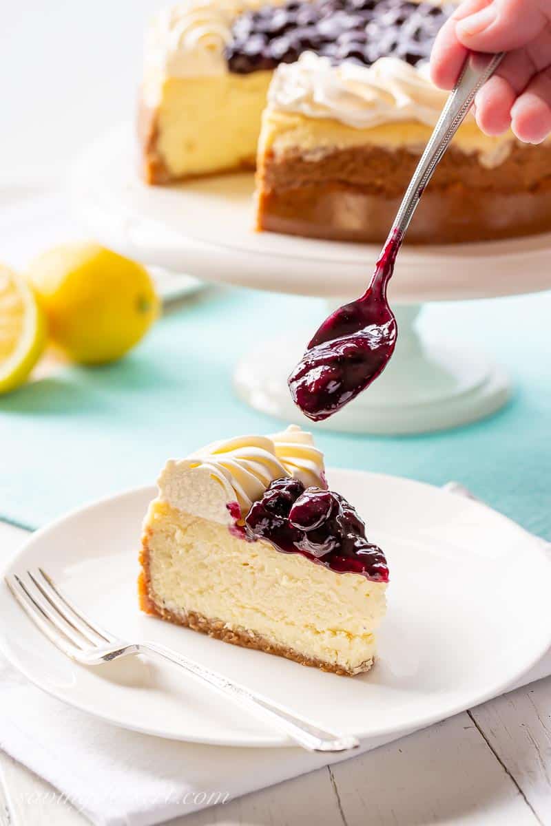 Blueberry sauce being spooned over a slice of cheesecake