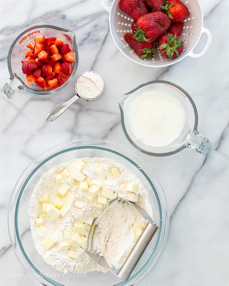 Bowls of ingredients on a marble slab with strawberries, flour, milk, butter and sugar