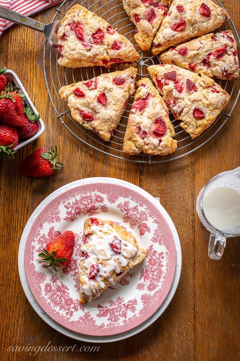 An overhead shot of a table with a scone on a plate, fresh strawberries, icing and a cooling rack with scone wedges