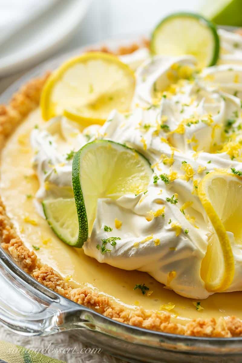 A side view of a lemon lime pie topped with whipped cream and sliced lemons and limes