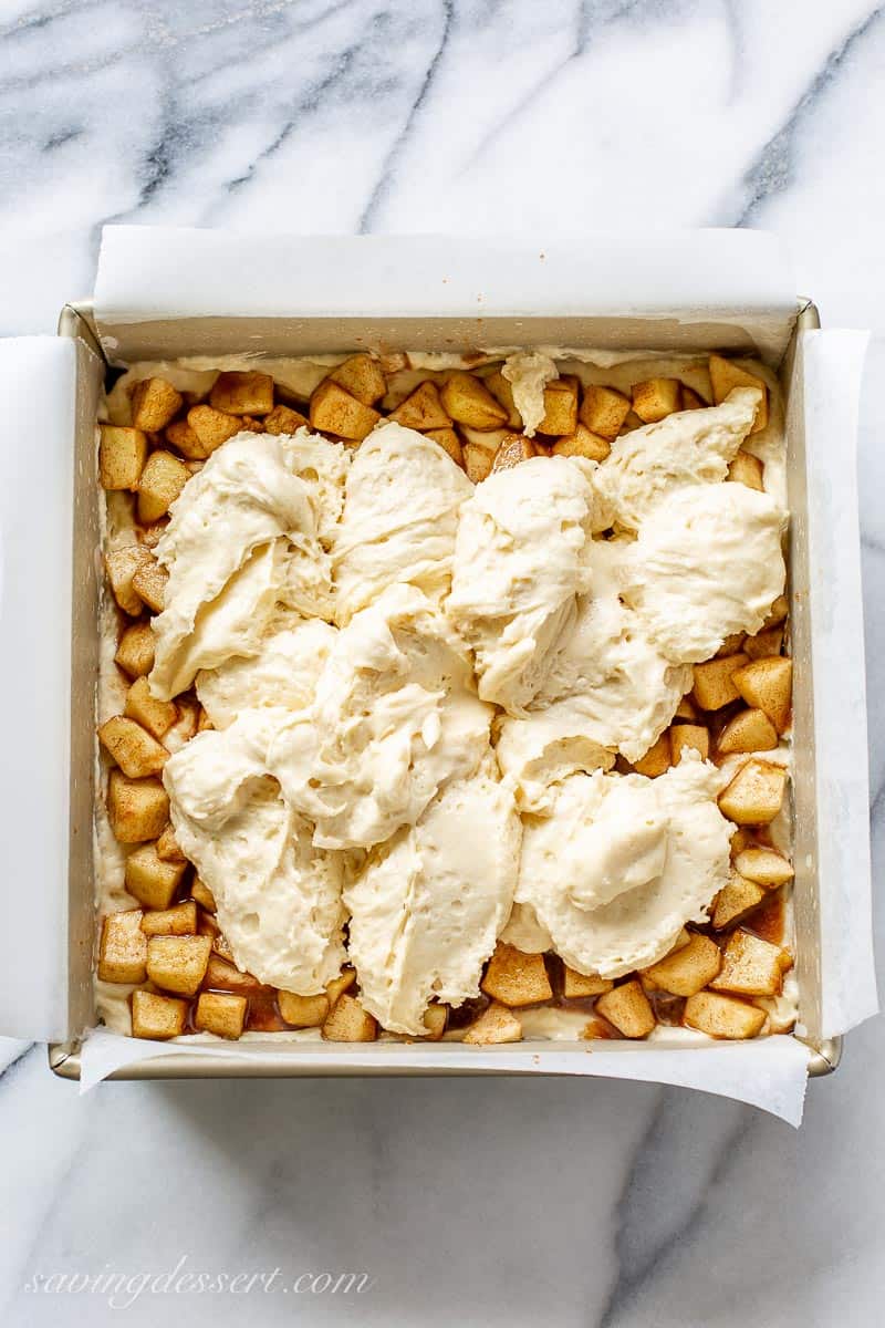 A square cake pan filled with cake batter, diced cinnamon apples and more cake batter dolloped on top