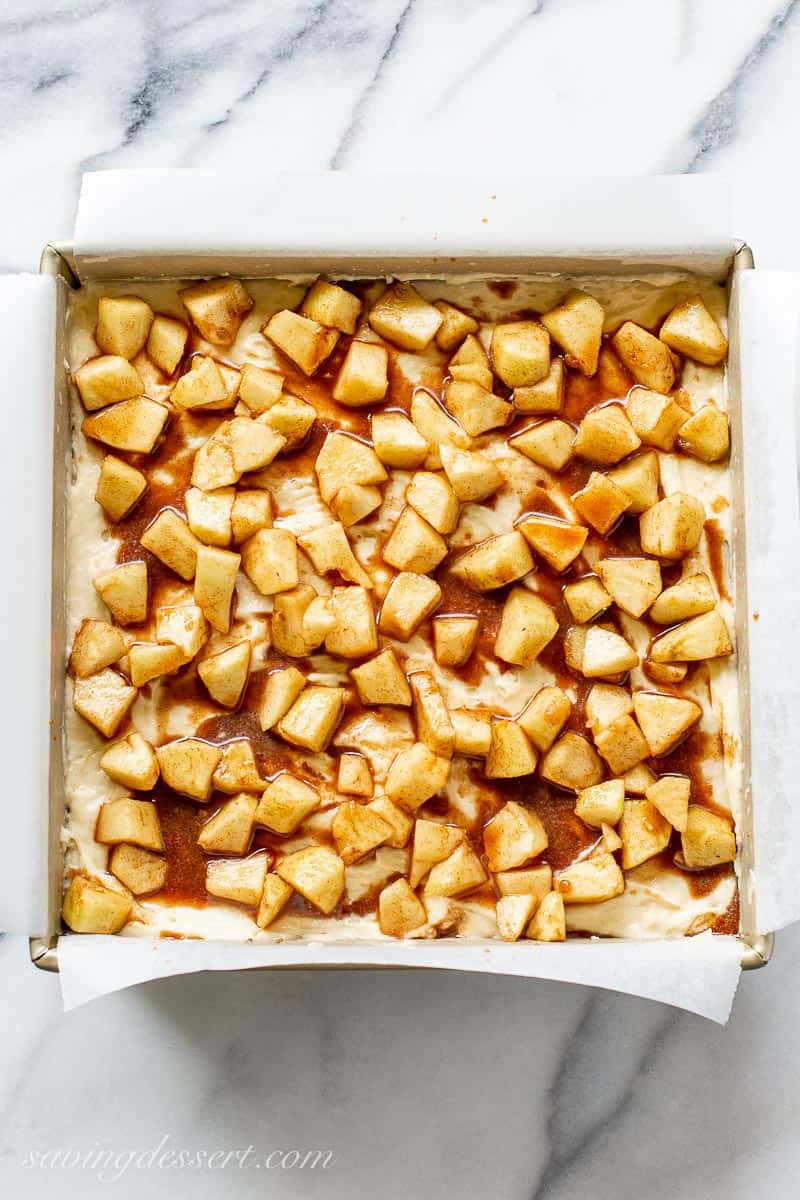 An overhead view of a square cake pan filled with thick cake batter topped with diced cinnamon apples