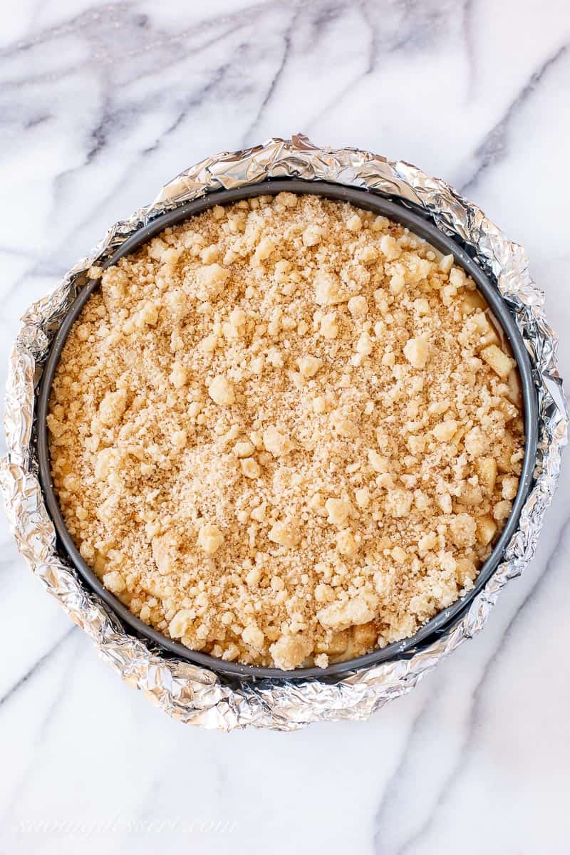 Overhead view of a foil wrapped springform pan filled with cheesecake batter and topped with diced apples and a crumble topping