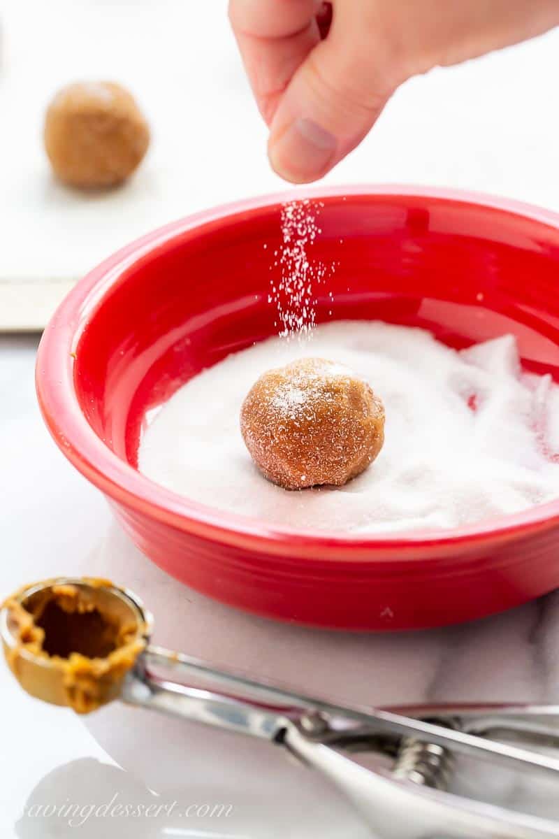 A ball of cookie dough in a bowl of granulated sugar with extra sugar being sprinkled on top