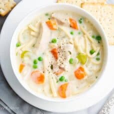 Overhead view of a bowl of creamy chicken noodle soup with carrots, peas and celery served with crackers