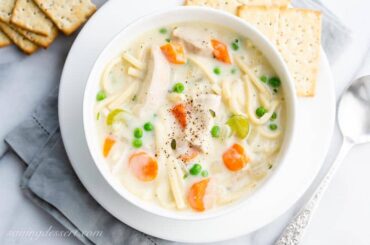 Overhead view of a bowl of creamy chicken noodle soup with carrots, peas and celery served with crackers