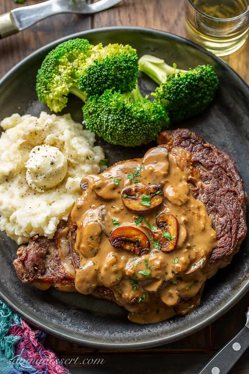 A steak on a plate covered in mushroom sauce and served with mashed potatoes and broccoli