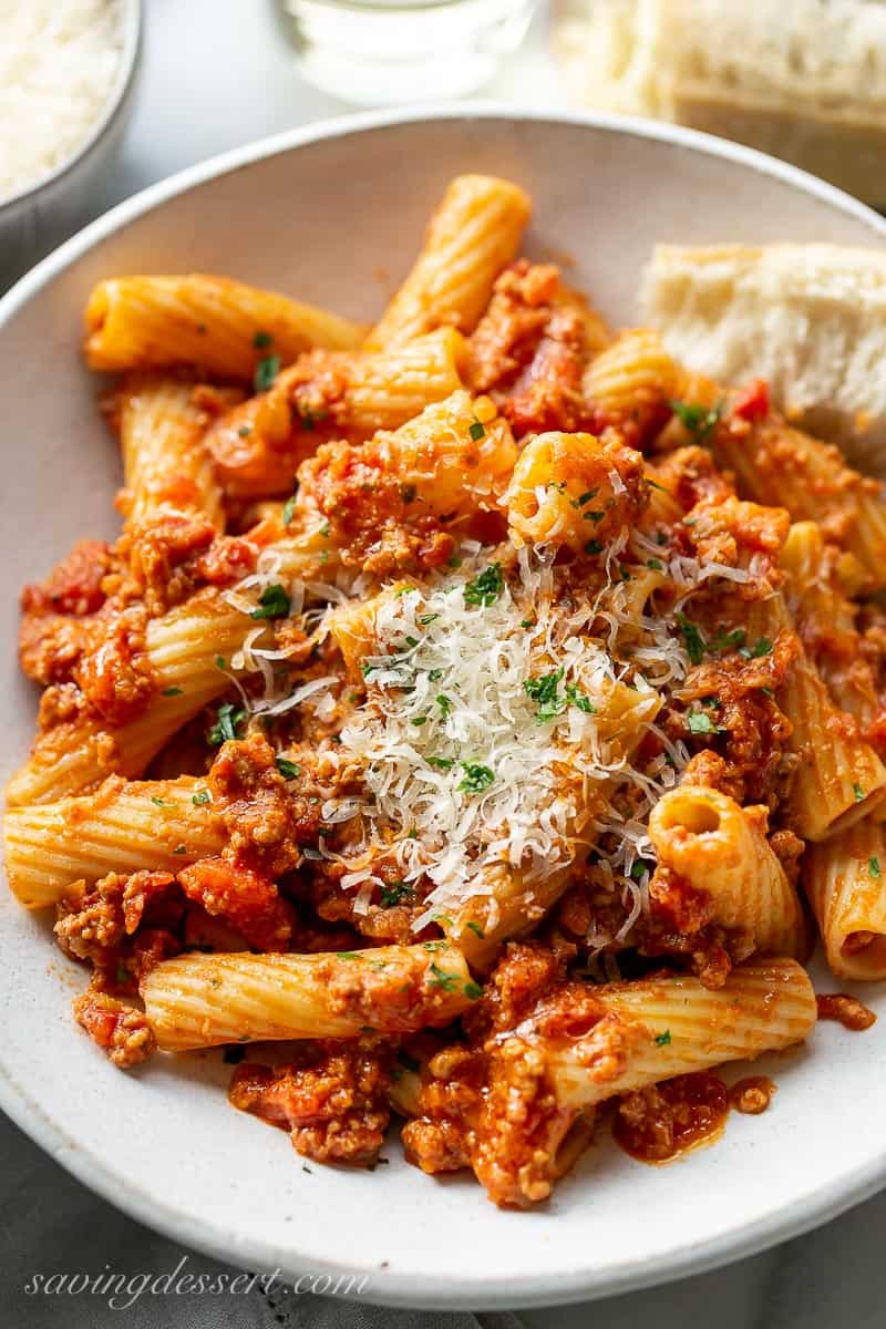 A bowl of meaty bolognese sauce with rigatoni pasta, crusty bread and fresh grated Parmesan