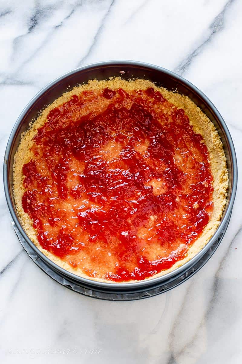 A golden Oreo crust in a springform pan topped with strawberry jam