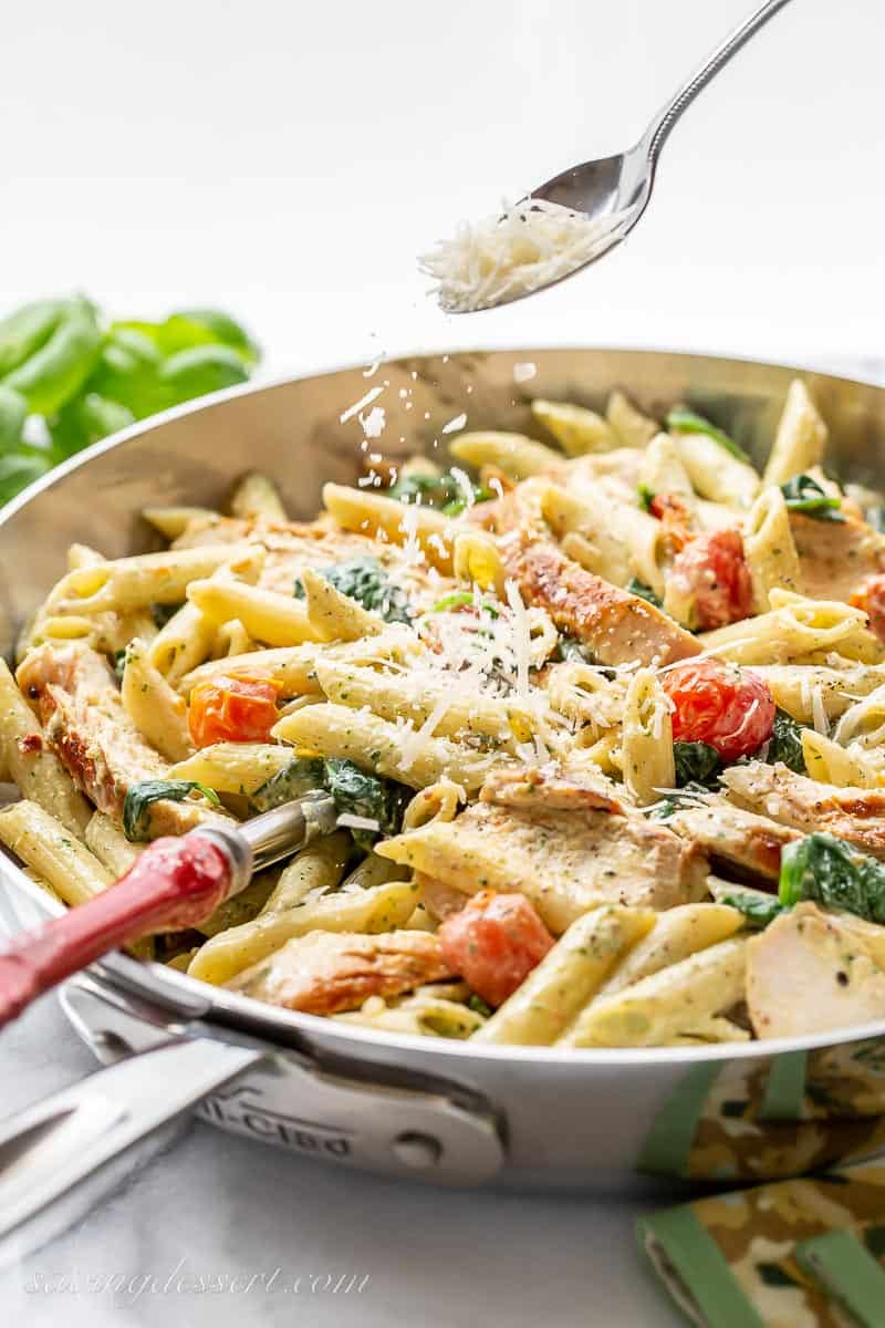 A side view of a skillet with chicken pesto pasta being sprinkled with fresh grated parmesan cheese