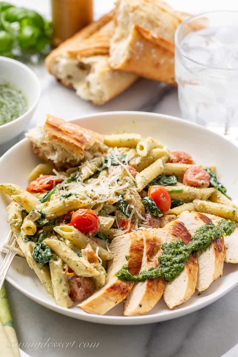 A bowl of chicken, pesto and pasta with a hunk of bread on the side