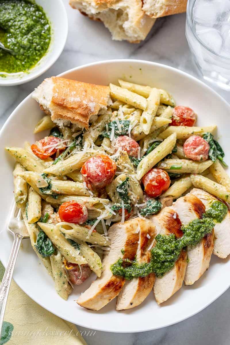 A bowl of pasta with sliced chicken, pesto sauce, tomatoes and a hunk of bread