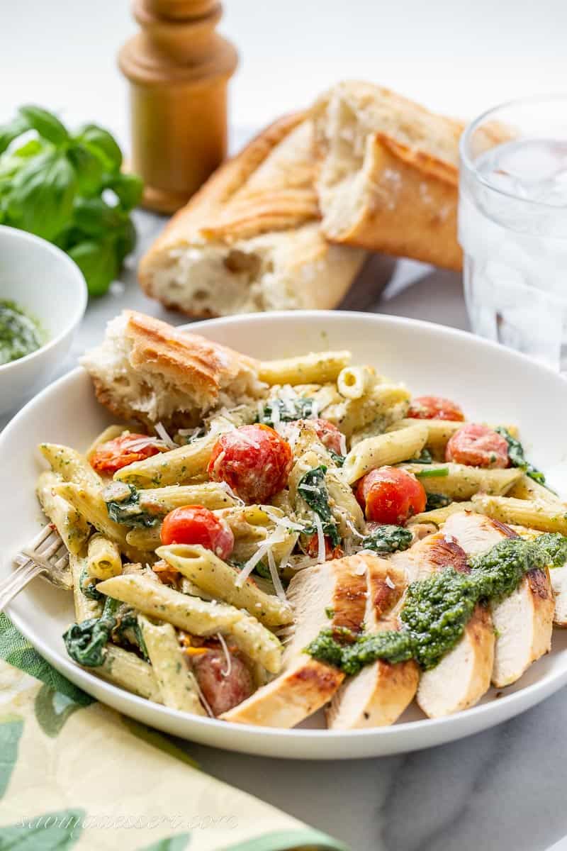 A bowl of pasta, sliced chicken, pesto sauce, tomatoes and a baguette