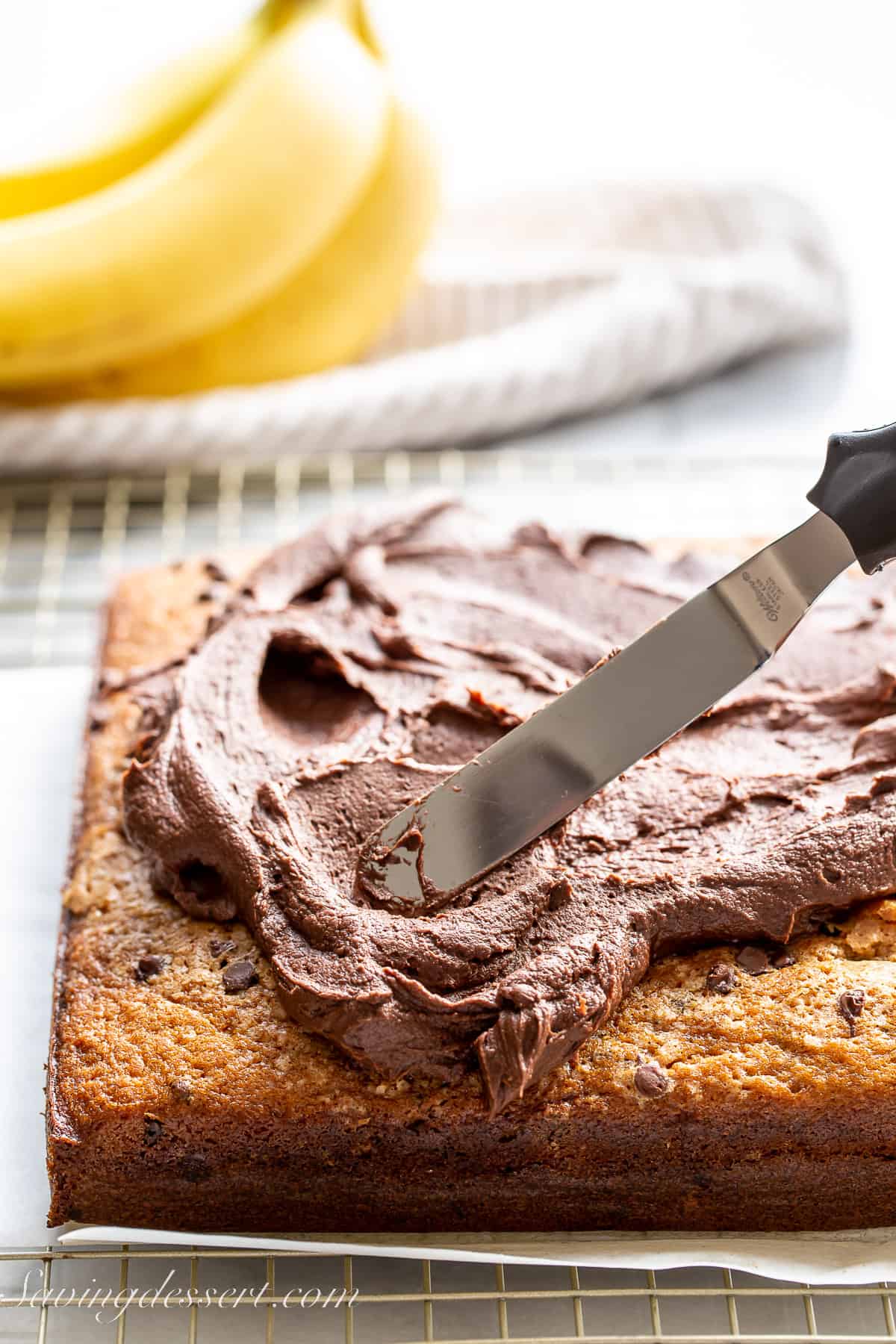 a square banana cake being iced with chocolate frosting