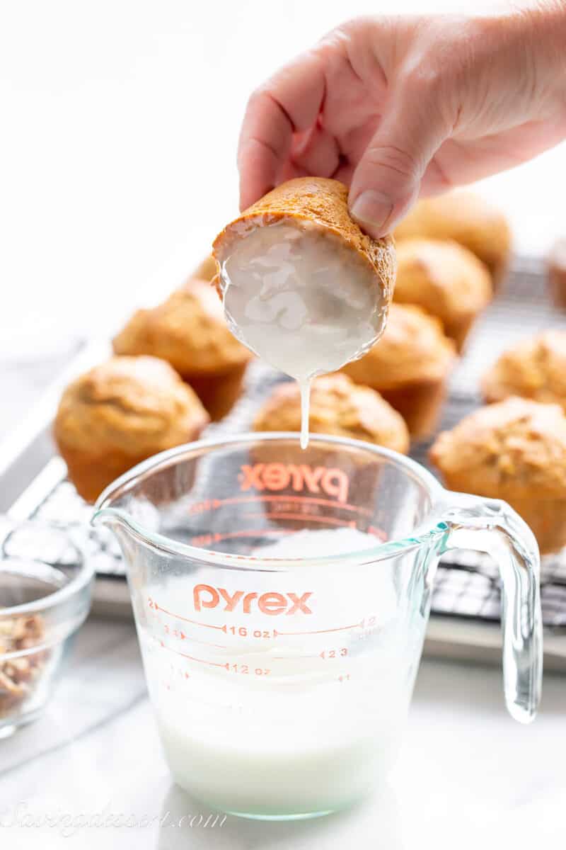 a muffin being dipped into a measuring cup filled with a powdered sugar glaze