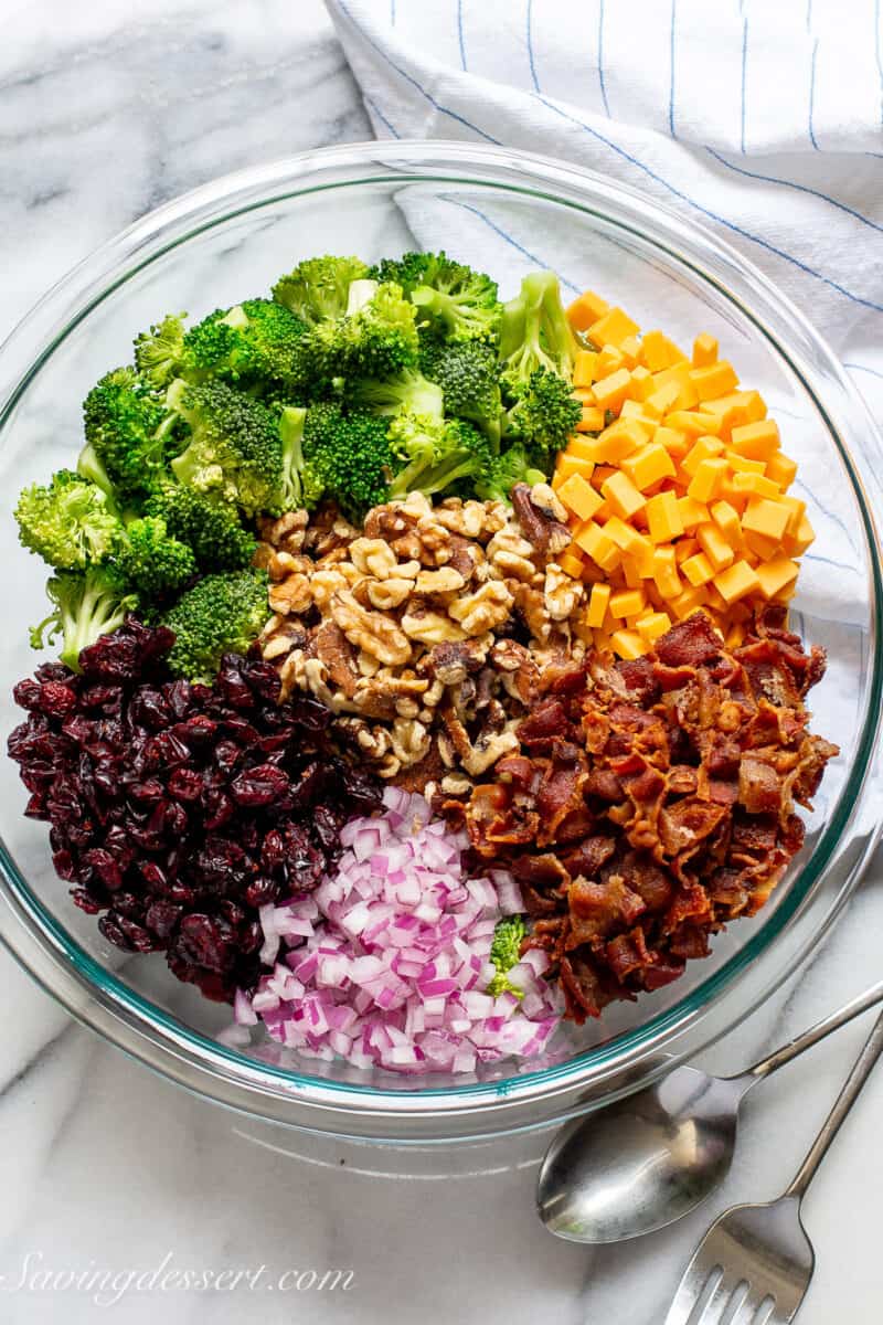 a large mixing bowl with piles of broccoli florets, walnuts, cheese squares, bacon, onion and craisins