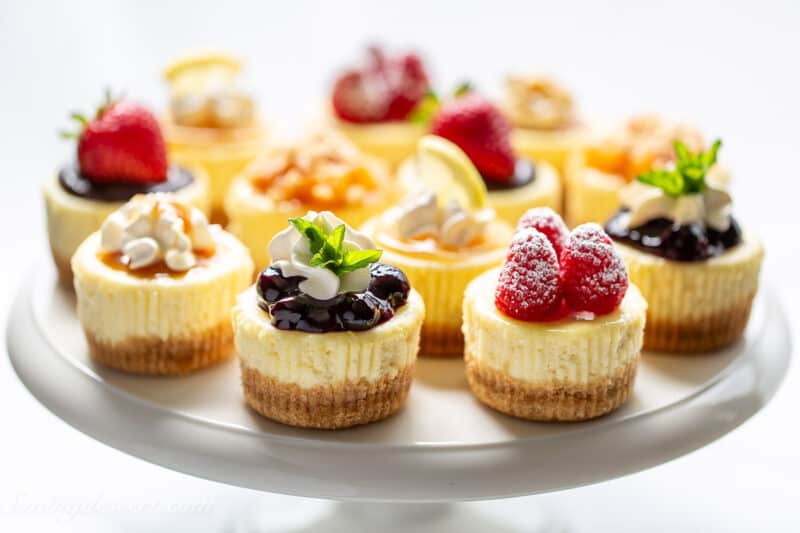 a cake plate covered with individual mini cheesecakes with various toppings