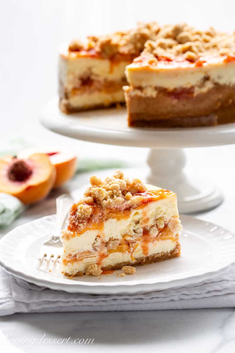 A slice of peach cobbler cheesecake on a plate with the remaining cake on a platter in the background.
