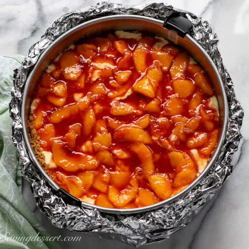A springform pan wrapped in foil with cheesecake batter and sliced peaches.