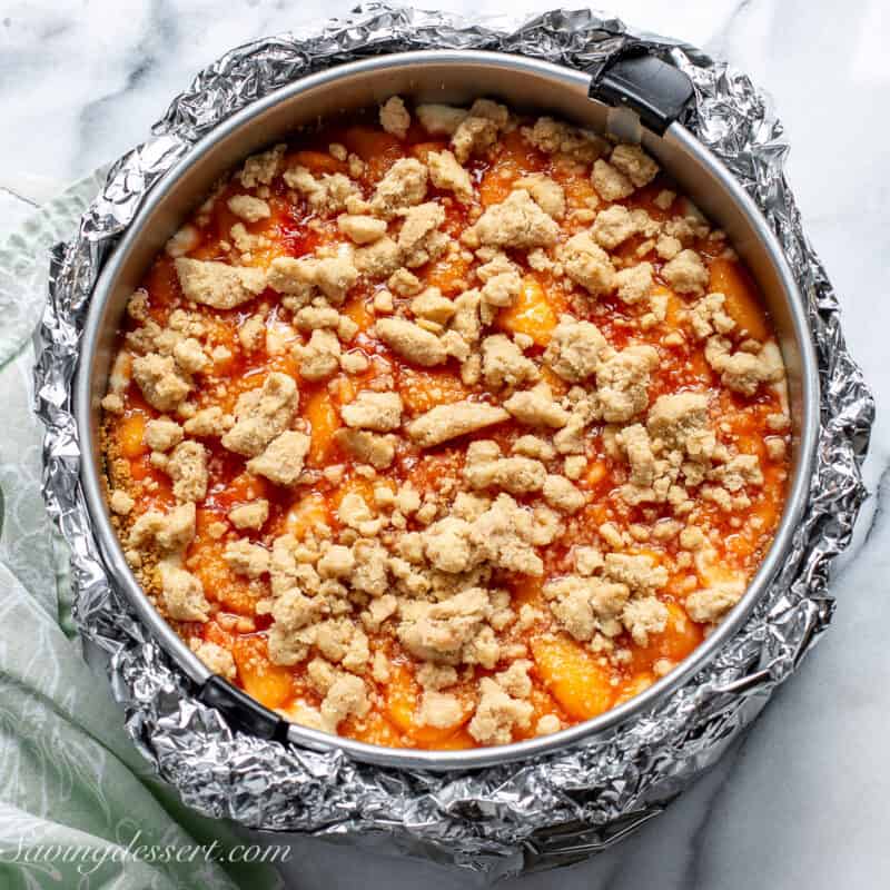 A springform pan wrapped in foil with a layer of peaches and a crumble topping.