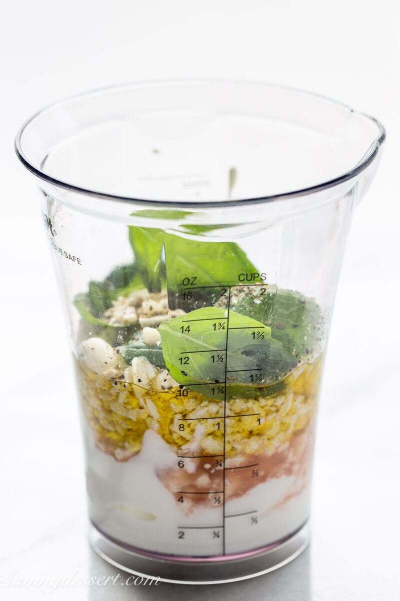 a measuring cup with ingredients for creamy feta dressing including fresh basil, feta cheese, olive oil and yogurt.