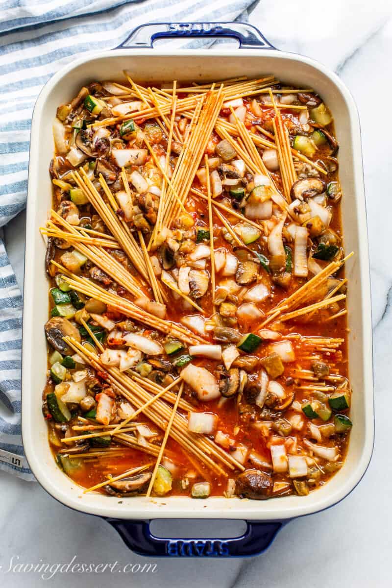 A casserole pan filled with vegetables, marinara and uncooked spaghetti noodles.