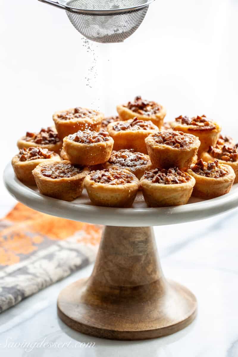 A cake stand filled with pecan tassies being sprinkled with powdered sugar.