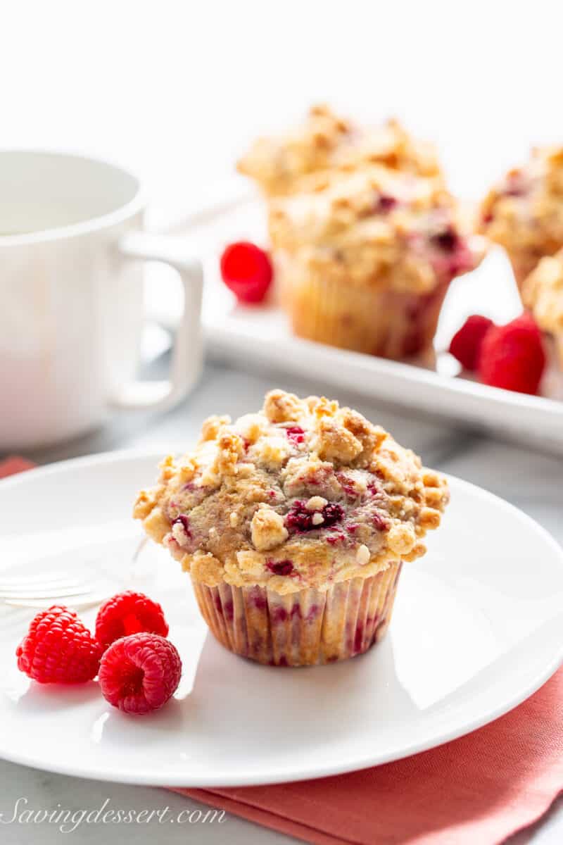 A plate with a raspberry muffin, fresh raspberries and a cup of coffee in the background.