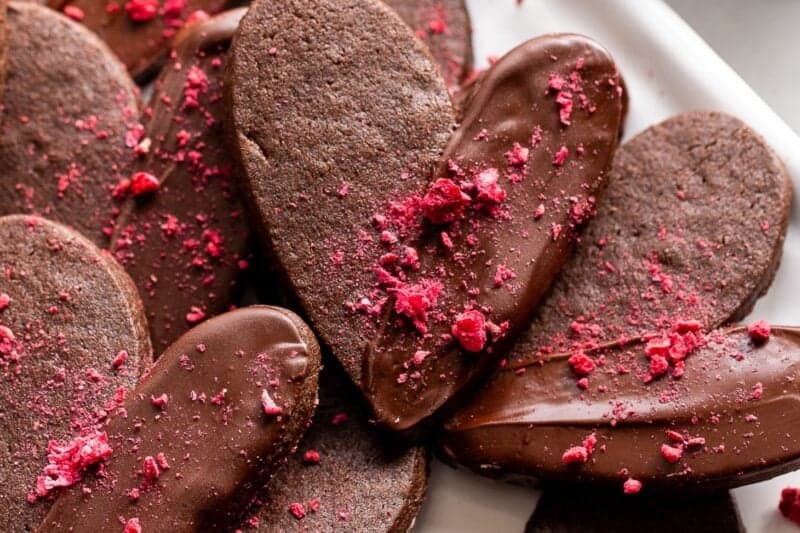 A closeup of a tray of chocolate heart shaped cookies dipped in dark chocolate and sprinkled with freeze dried raspberries.
