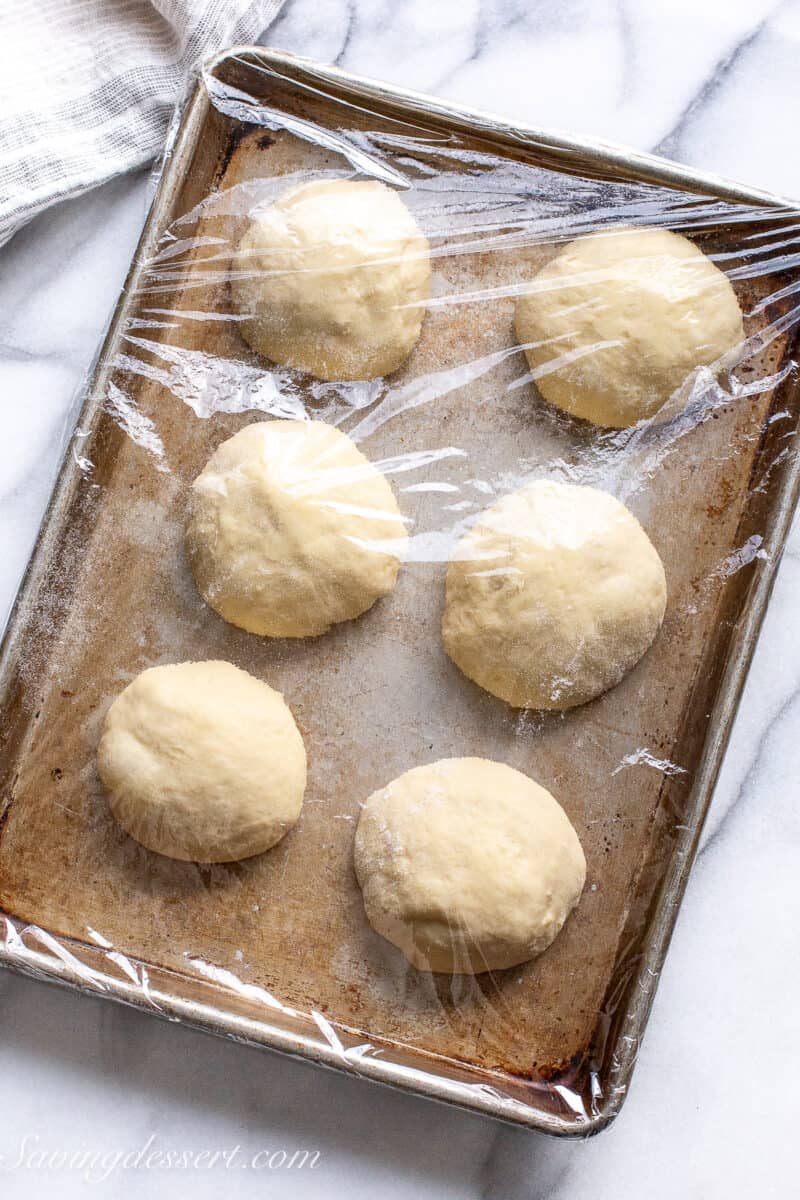 A small baking tray with 6 dough balls spaced across the pan which is covered with plastic wrap.