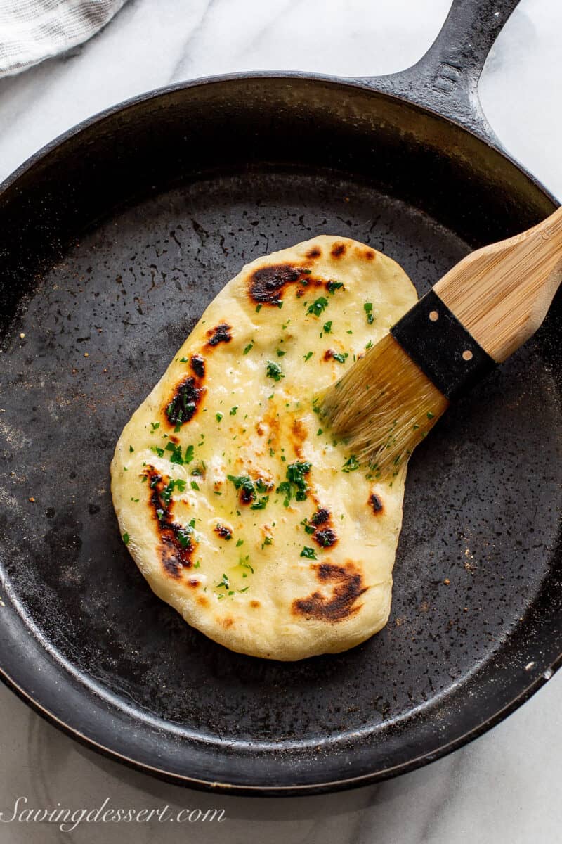 A naan bread round in a cast iron skillet being brushed with melted parsley butter.