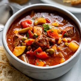 A closeup photo of a bowl of hearty vegetable beef soup with bread on the side.