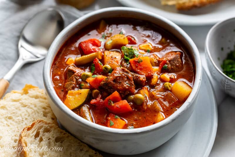 A closeup photo of a bowl of hearty vegetable beef soup with bread on the side.