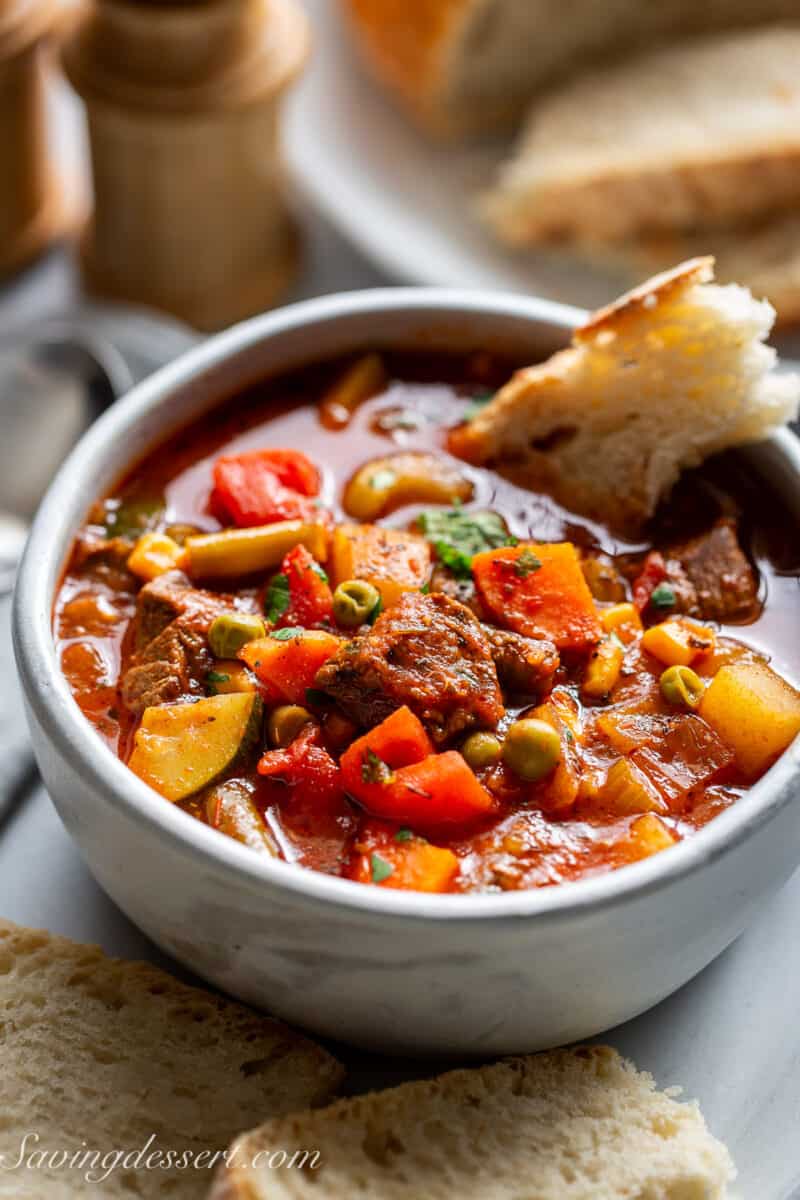 A big bowl of hearty vegetable beef soup in a bowl with a piece of crusty bread dipped in.