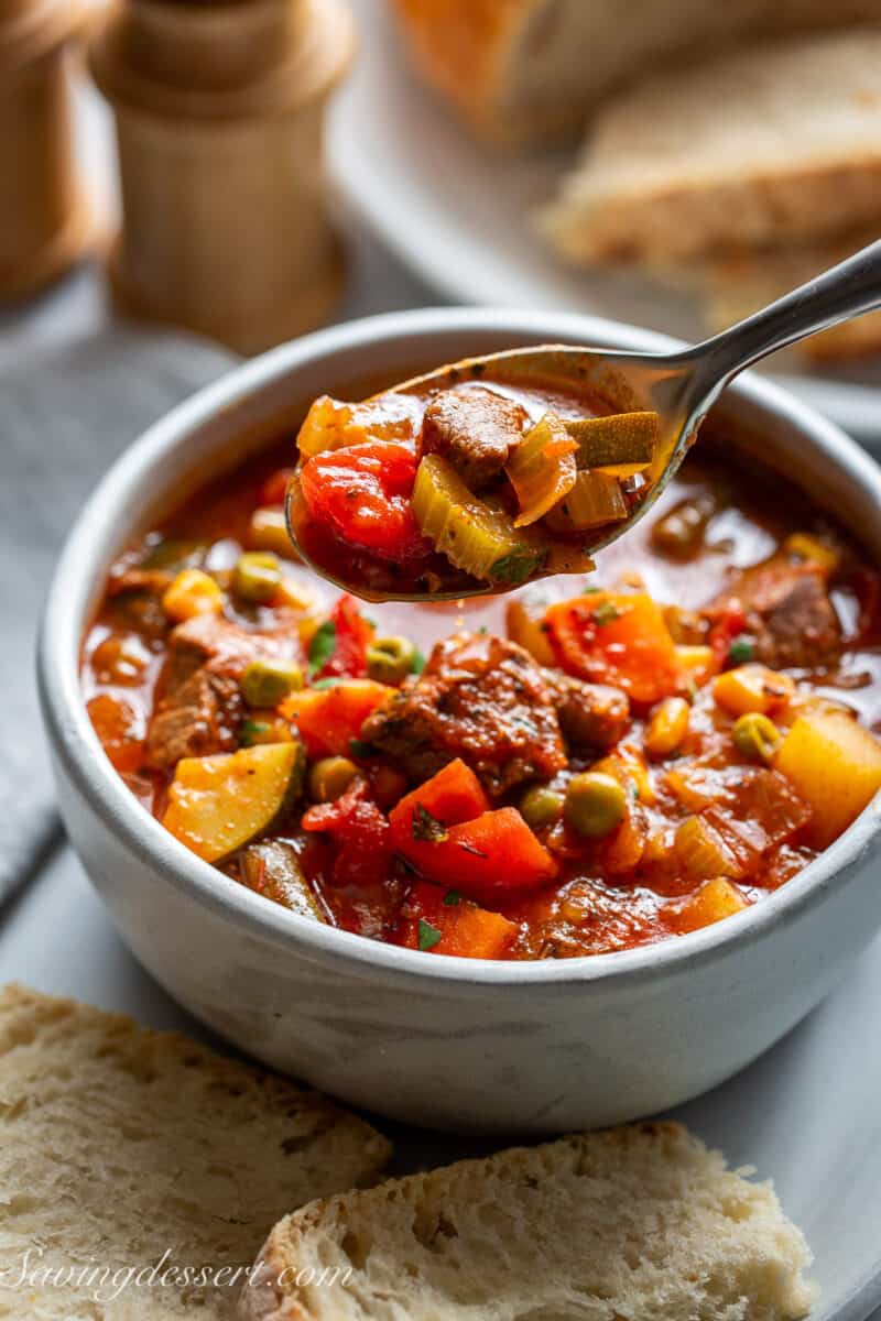 A bowl of vegetable beef soup with a spoonful being removed with sliced bread on the side.