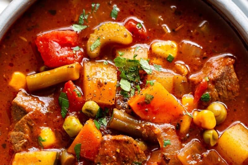An overhead bowl of vegetable beef soup with chunky pieces of zucchini, tomatoes, potatoes, beef, peaches and carrots.