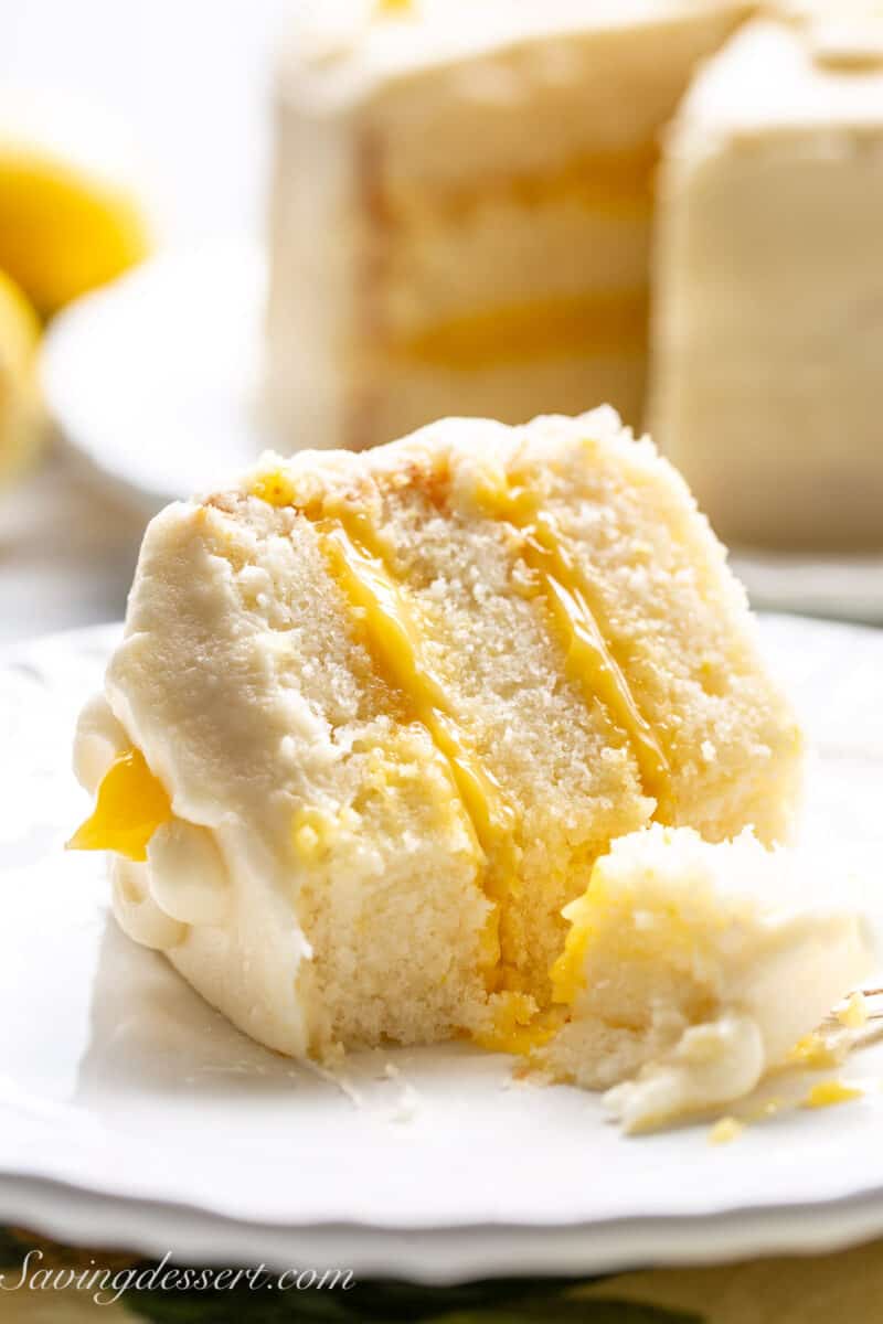 A closeup of a partially eaten slice of lemon curd cake on a plate with the remaining cake in the background.