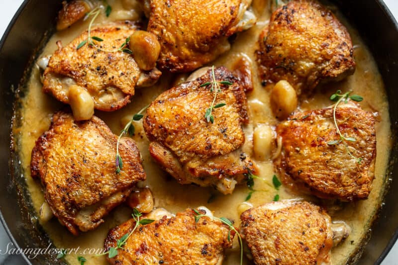 Chicken thighs in a skillet filled with 40 cloves of garlic in a buttery wine sauce.