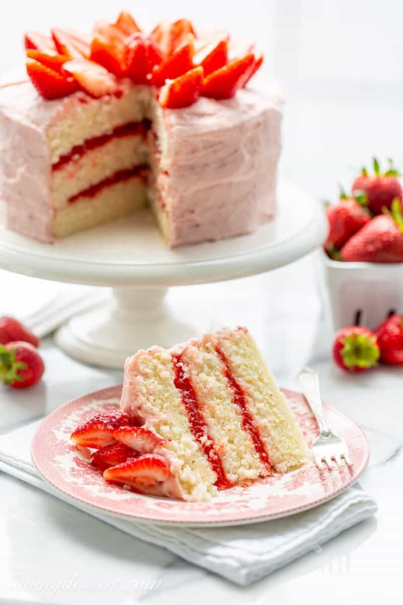 A slice of strawberry lemonade cake with the whole cake on a cake stand in the background.